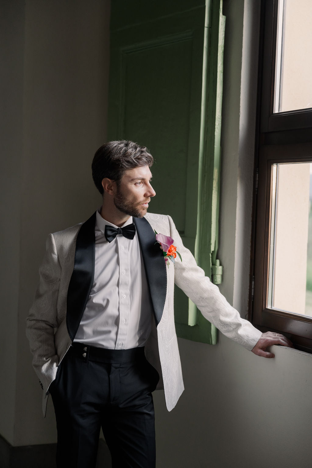 Man in a formal suit standing by a window with a decorative flower boutonniere.