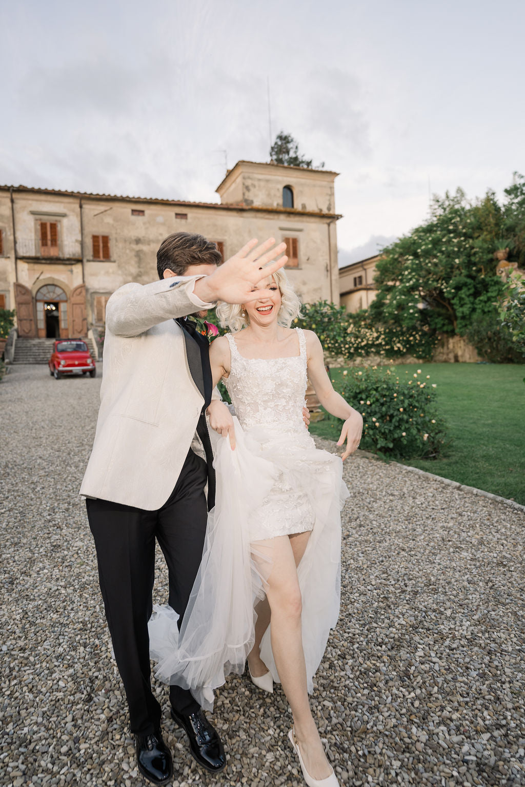 Man and woman in formal attire walking outdoors with a building and greenery in the background