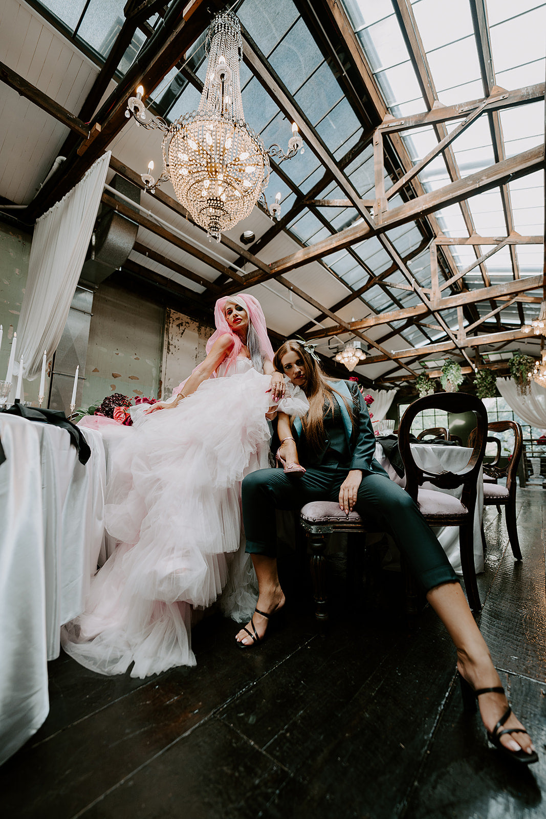 Two women in formal attire sitting together in a stylish indoor setting with a chandelier.